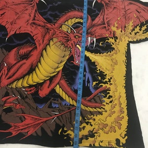 May include: Black t-shirt featuring a large red dragon with yellow accents, spewing flames. The dragon has large wings and sharp teeth. A blue measuring tape is placed on the shirt, showing the length in inches.