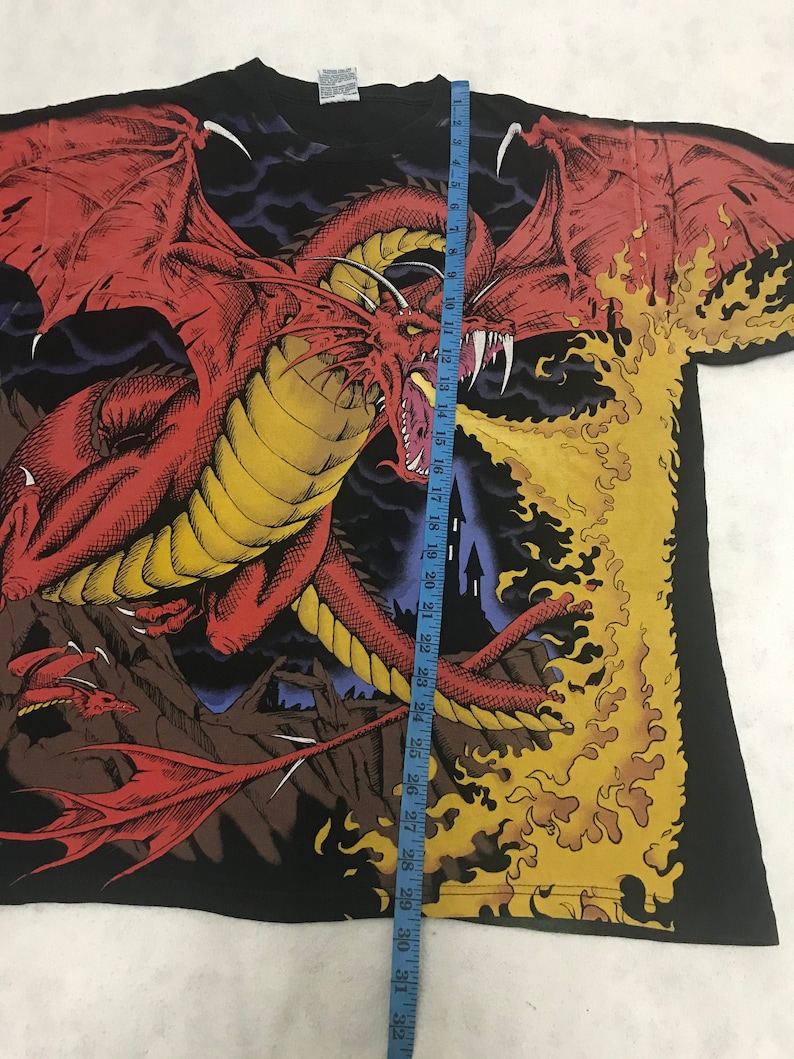 May include: Black t-shirt featuring a large red dragon with yellow accents, spewing flames. The dragon has large wings and sharp teeth. A blue measuring tape is placed on the shirt, showing the length in inches.