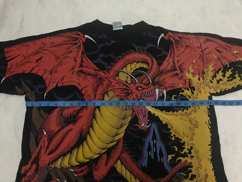 May include: Black t-shirt featuring a detailed red dragon graphic with large wings, spewing flames. The dragon is set against a dark background with a castle silhouette. The shirt's design covers the entire front.