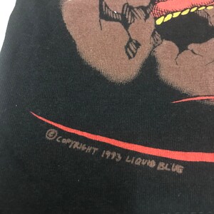 May include: Black t-shirt featuring a red and brown graphic design of a creature. The graphic is detailed with sharp claws and teeth. The text "COPYRIGHT 1993 LIQUID BLUE" is printed in brown along the bottom edge of the shirt.