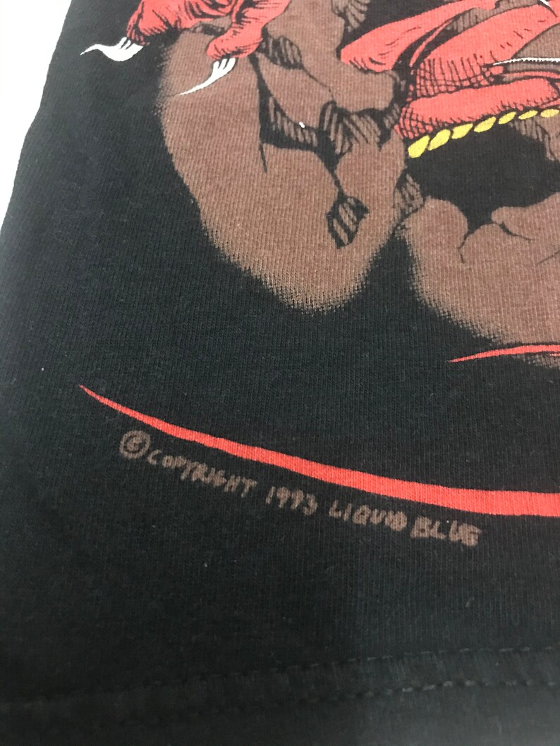 May include: Black t-shirt featuring a red and brown graphic design of a creature. The graphic is detailed with sharp claws and teeth. The text "COPYRIGHT 1993 LIQUID BLUE" is printed in brown along the bottom edge of the shirt.