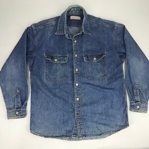 Puede incluir: Una camisa vaquera azul con mangas largas, cierre de botones y dos bolsillos en el pecho. La camisa tiene un cuello clásico y puños abotonados. La marca "DEAVAN" es visible en la etiqueta. La camisa está tendida sobre una superficie blanca.