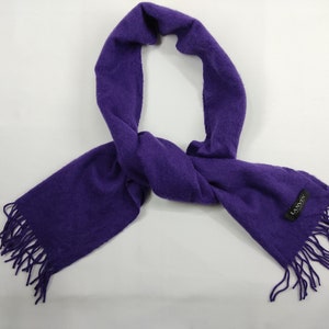 Puede incluir: Una bufanda de color púrpura vibrante con bordes con flecos. La bufanda está hecha de un material suave y texturizado y presenta una pequeña etiqueta negra con texto blanco. La bufanda se muestra sobre un fondo blanco.