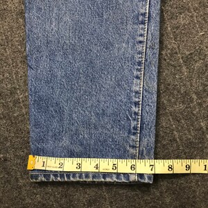 May include: Close-up of blue denim jeans with a yellow measuring tape. The tape shows a measurement of approximately 15 cm. The jeans have a classic, faded wash and visible stitching.