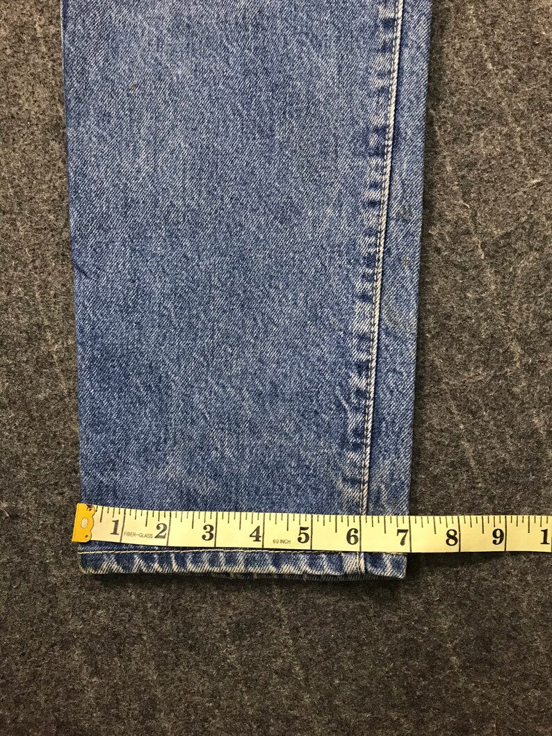 May include: Close-up of blue denim jeans with a yellow measuring tape. The tape shows a measurement of approximately 15 cm. The jeans have a classic, faded wash and visible stitching.