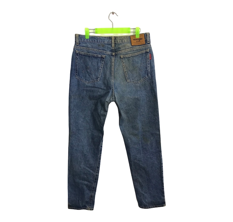 May include: Blue denim Wrangler jeans with a classic five-pocket design. The jeans are hanging on a green hanger, showcasing the back pockets and the Wrangler brand patch. The denim has a faded wash.