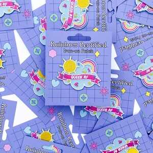 May include: Iron-on patches with a rainbow, sun, and cloud design, with the text "QUEER AF" on a pink banner. The patches are on purple cards that read "Rainbow Certified Iron-on Patch".