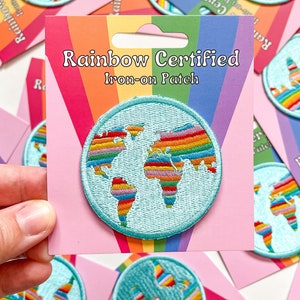 Op de afbeelding: Een ronde, lichtblauwe opstrijkbare patch met een regenboogkleurig wereldkaartontwerp. De patch is gelabeld met "Rainbow Certified" op een roze en regenboog gestreepte achtergrondkaart. Meerdere patches zijn zichtbaar.
