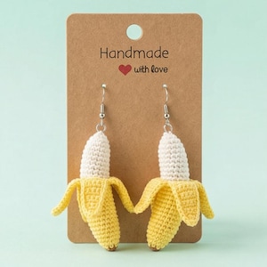 May include: A pair of handmade crochet banana earrings. The earrings feature a yellow banana peel with a white banana inside. The earrings are hanging on a brown card that says "Handmade with love".