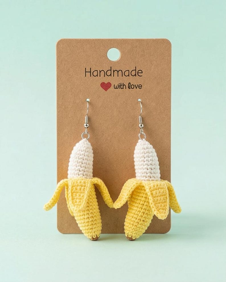 May include: A pair of handmade crochet banana earrings. The earrings feature a yellow banana peel with a white banana inside. The earrings are hanging on a brown card that says "Handmade with love".