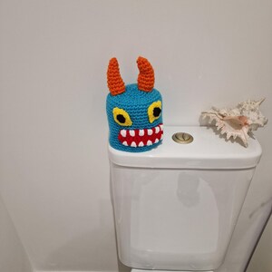Puede incluir: Una funda de asiento de inodoro de monstruo de ganchillo azul con cuernos naranjas, ojos amarillos y boca roja. La funda hecha a mano se coloca en un inodoro blanco, agregando un toque caprichoso a la decoraci&oacute;n del ba&ntilde;o.
