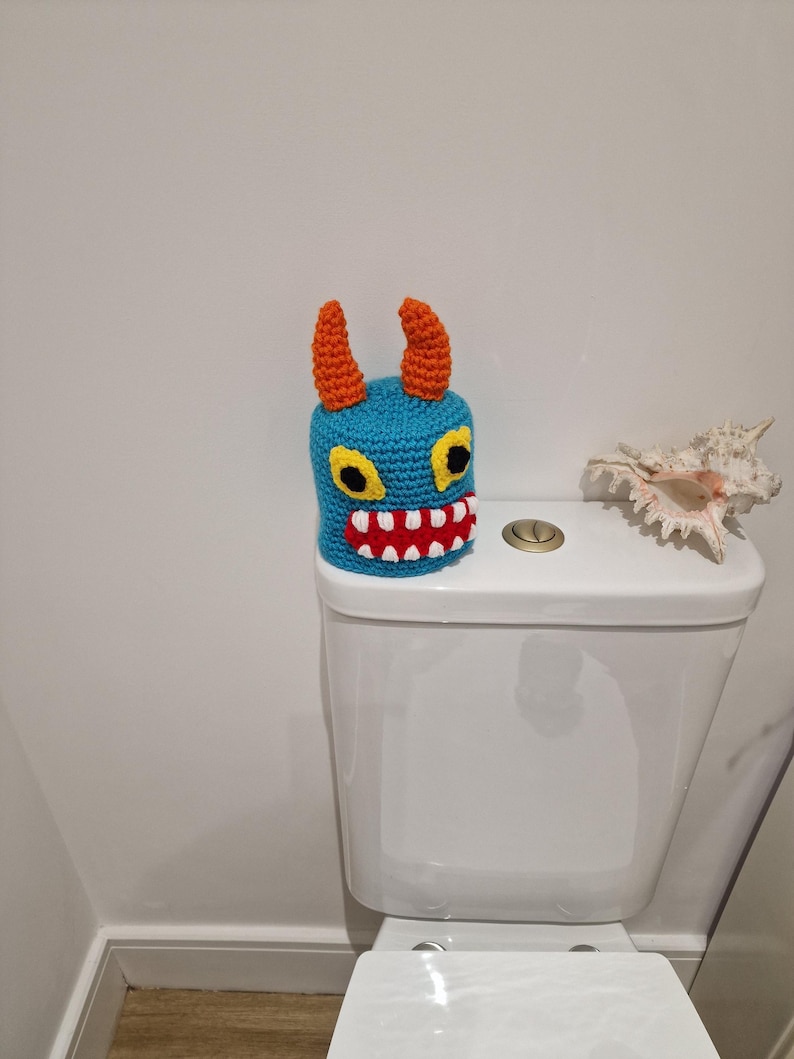 Puede incluir: Una funda de asiento de inodoro de monstruo de ganchillo azul con cuernos naranjas, ojos amarillos y boca roja. La funda hecha a mano se coloca en un inodoro blanco, agregando un toque caprichoso a la decoraci&oacute;n del ba&ntilde;o.