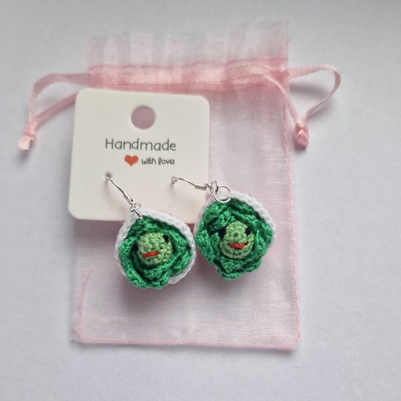 K&ouml;nnte beinhalten: Ein Paar handgefertigte geh&auml;kelte Kohl-Ohrringe mit gr&uuml;nen Bl&auml;ttern und einem roten Mund. Die Ohrringe befinden sich auf einer wei&szlig;en Karte mit dem Text "Handmade with love".