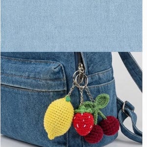 Peut inclure: Un sac &agrave; dos en jean bleu avec une poche avant et une fermeture &eacute;clair argent&eacute;e. Le sac &agrave; dos est orn&eacute; d'un porte-cl&eacute;s avec des breloques de fruits crochet&eacute;s : un citron jaune, une fraise rouge et deux cerises marron.