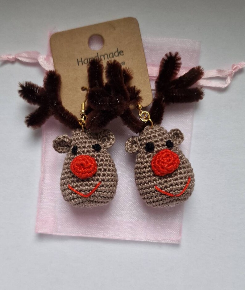 K&ouml;nnte beinhalten: Ein Paar handgefertigte geh&auml;kelte Rentierohrringe mit braunen Geweihen und roten Nasen. Die Ohrringe sind in einem rosa Organzas&auml;ckchen mit einem braunen Etikett verpackt, auf dem "Handmade" steht.