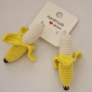 May include: A pair of handmade crochet banana earrings. The earrings feature a yellow peel and a white banana with a black tip. The earrings are hanging from a white tag that says "Handmade with love".