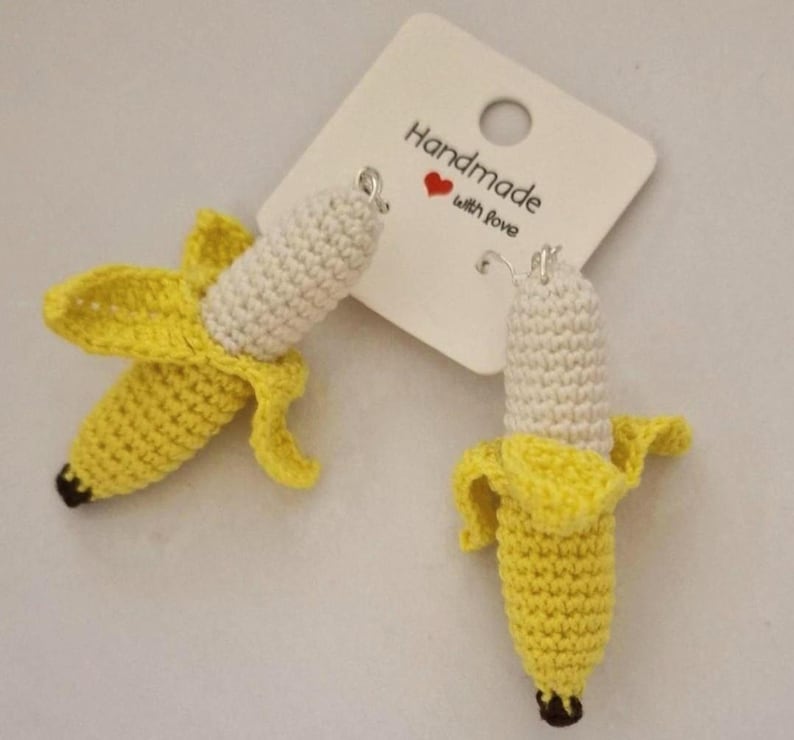 May include: A pair of handmade crochet banana earrings. The earrings feature a yellow peel and a white banana with a black tip. The earrings are hanging from a white tag that says "Handmade with love".