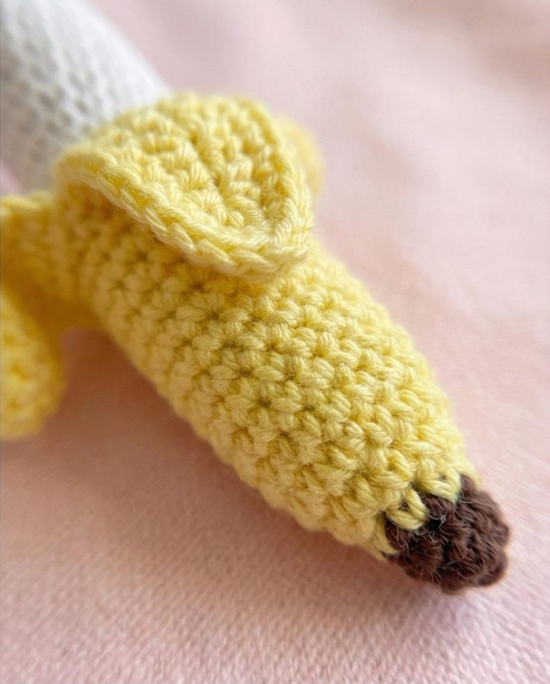May include: A handmade crochet banana in shades of yellow and brown. The banana peel is a bright yellow, with a textured surface. The tip of the banana is brown. The banana is partially peeled, revealing a white interior.