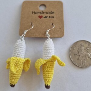 May include: Handmade crochet banana earrings with silver hooks. The earrings feature a yellow banana peel and a white banana body, with a brown tip. The earrings are displayed with a tag that reads "Handmade with love".