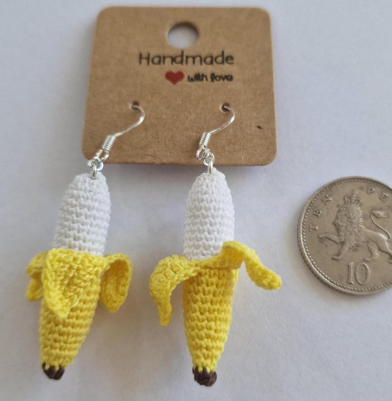 May include: Handmade crochet banana earrings with silver hooks. The earrings feature a yellow banana peel and a white banana body, with a brown tip. The earrings are displayed with a tag that reads "Handmade with love".