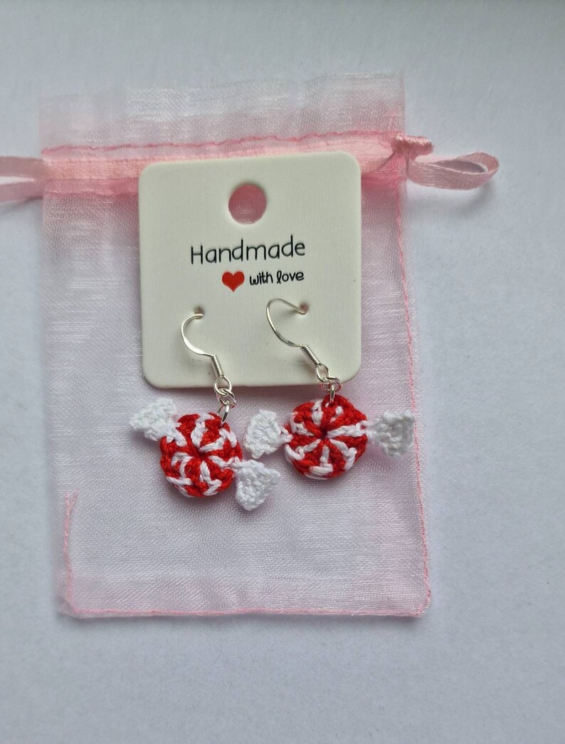 K&ouml;nnte beinhalten: Geh&auml;kelte Zuckerstangen-Ohrringe mit silbernen Haken. Die Ohrringe sind in einem rosa Organzas&auml;ckchen mit einem wei&szlig;en Etikett verpackt, auf dem "Handmade with love" steht.