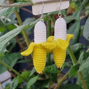 Crochet banana earrings.  A pair of yellow and white crocheted banana dangle earrings with silver ear wires.  The earrings are displayed on a white card with the text 'Handmade with Love'. The background shows green foliage.