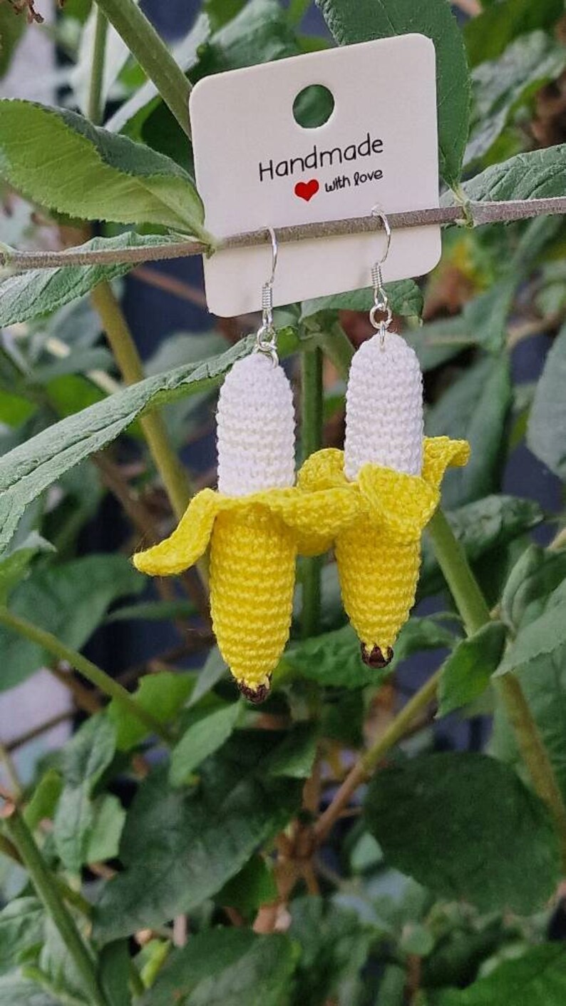 Crochet banana earrings.  A pair of yellow and white crocheted banana dangle earrings with silver ear wires.  The earrings are displayed on a white card with the text 'Handmade with Love'. The background shows green foliage.