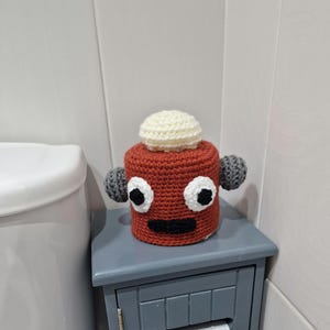 Puede incluir: Un portarrollos de papel higi&eacute;nico de ganchillo hecho a mano con forma de robot de dibujos animados. Presenta grandes ojos blancos y negros, una boca negra, una cabeza color crema y ap&eacute;ndices grises en forma de orejas. El soporte se encuentra sobre un peque&ntilde;o armario gris.