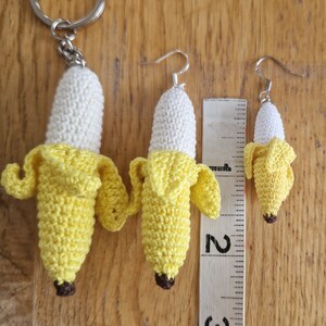 Three crocheted banana keychain and earrings in varying sizes. The largest banana is attached to a silver key ring, while the smaller bananas are fashioned as earrings with silver hooks.  All bananas feature a white body and yellow peel, with a brown stem detail. A ruler is included for size comparison, showing measurements 2 and 3.