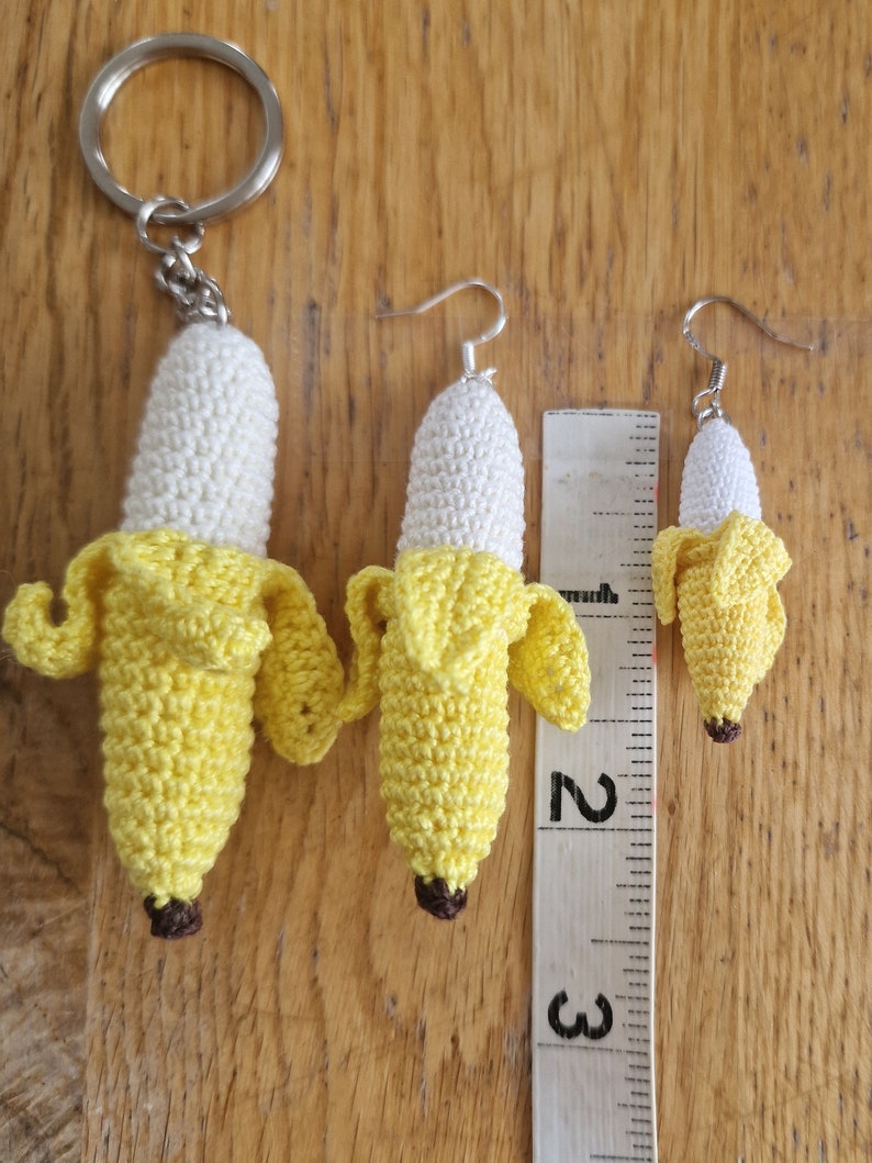 Three crocheted banana keychain and earrings in varying sizes. The largest banana is attached to a silver key ring, while the smaller bananas are fashioned as earrings with silver hooks.  All bananas feature a white body and yellow peel, with a brown stem detail. A ruler is included for size comparison, showing measurements 2 and 3.