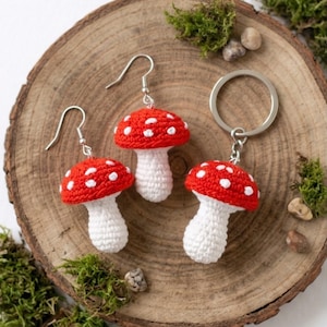 May include: Crocheted mushroom earrings and keychain set. The earrings and keychain feature red caps with white spots and white stems. The items are displayed on a wooden slice with moss and small stones.