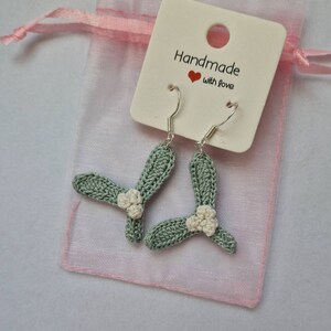 K&ouml;nnte beinhalten: Ein Paar gr&uuml;ne H&auml;kelohrringe mit wei&szlig;en Blumenakzenten. Die Ohrringe h&auml;ngen an silbernen Haken und sind in einem rosa Organzas&auml;ckchen mit einem wei&szlig;en Etikett verpackt, auf dem "Handmade with love" steht.