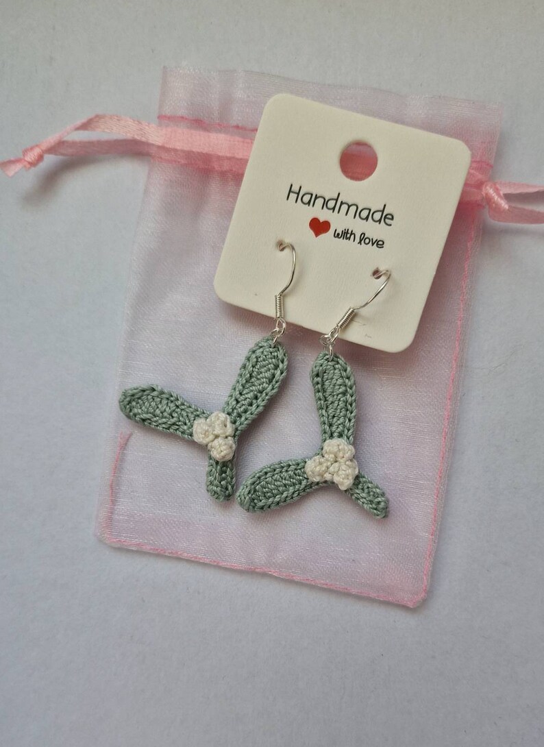 K&ouml;nnte beinhalten: Ein Paar gr&uuml;ne H&auml;kelohrringe mit wei&szlig;en Blumenakzenten. Die Ohrringe h&auml;ngen an silbernen Haken und sind in einem rosa Organzas&auml;ckchen mit einem wei&szlig;en Etikett verpackt, auf dem "Handmade with love" steht.