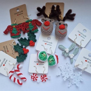 K&ouml;nnte beinhalten: Eine Auswahl an handgefertigten Ohrringen zum Thema Weihnachten. Die Ohrringe sind aus geh&auml;keltem Garn gefertigt und zeigen verschiedene Designs, darunter Rentiere, Stechpalmen, Zuckerstangen, Schneeflocken und Weihnachtspullover. Die Ohrringe werden auf wei&szlig;en Karten mit dem Text "Handmade with love" pr&auml;sentiert.