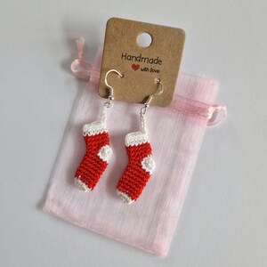 K&ouml;nnte beinhalten: Ein Paar rote und wei&szlig;e geh&auml;kelte Weihnachtsstrumpf-Ohrringe mit silbernen Haken. Die Ohrringe sind in einem rosa Organzas&auml;ckchen verpackt mit einem braunen Etikett, auf dem "Handmade with love" steht.