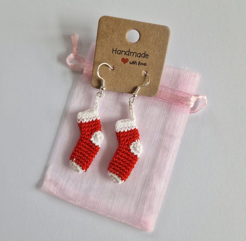 K&ouml;nnte beinhalten: Ein Paar rote und wei&szlig;e geh&auml;kelte Weihnachtsstrumpf-Ohrringe mit silbernen Haken. Die Ohrringe sind in einem rosa Organzas&auml;ckchen verpackt mit einem braunen Etikett, auf dem "Handmade with love" steht.