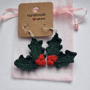 K&ouml;nnte beinhalten: Ein Paar gr&uuml;ne und rote geh&auml;kelte Ohrringe in Form von Stechpalmenbl&auml;ttern mit silbernen Haken. Die Ohrringe sind in einem rosa Organzas&auml;ckchen mit einem braunen Etikett verpackt, auf dem "Handmade with love" steht.