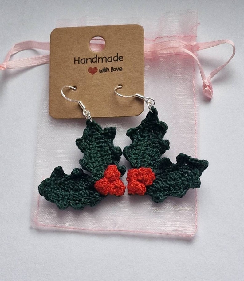 K&ouml;nnte beinhalten: Ein Paar gr&uuml;ne und rote geh&auml;kelte Ohrringe in Form von Stechpalmenbl&auml;ttern mit silbernen Haken. Die Ohrringe sind in einem rosa Organzas&auml;ckchen mit einem braunen Etikett verpackt, auf dem "Handmade with love" steht.
