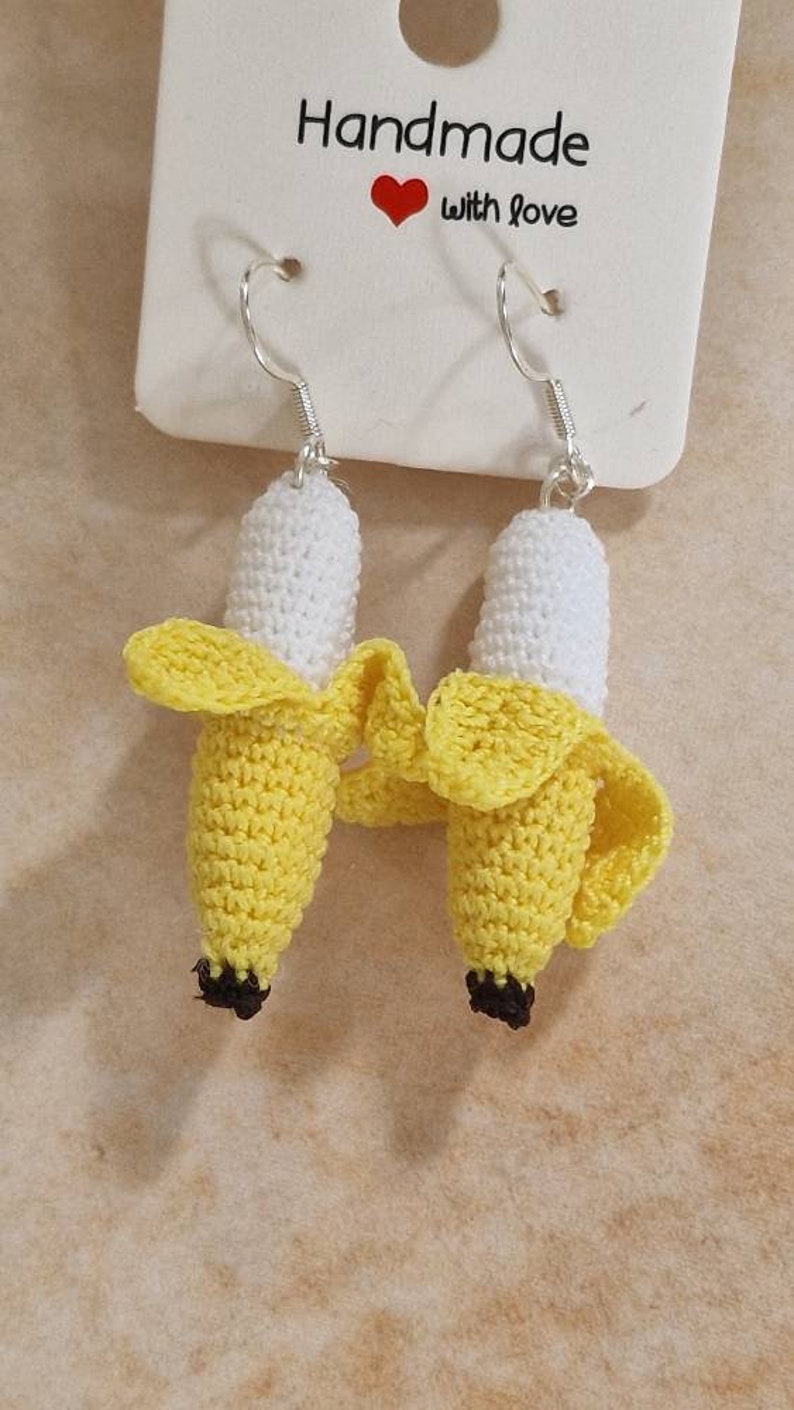 Handmade crocheted banana earrings.  The earrings are yellow and white, with silver hooks. The packaging shows the words 'Handmade with love'. These charming, whimsical earrings are perfect for adding a touch of fun to any outfit.