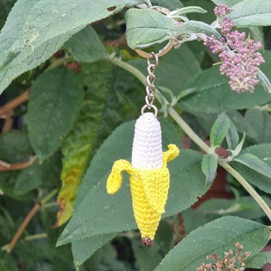 A crocheted banana keychain. The banana is predominantly yellow with a white center. It features a detailed peel and is attached to a silver metal key ring. The keychain is shown hanging from a green leafy plant.