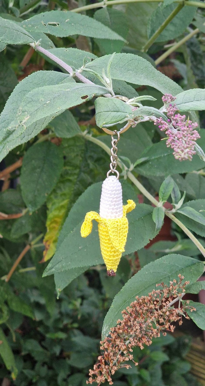 A crocheted banana keychain. The banana is predominantly yellow with a white center. It features a detailed peel and is attached to a silver metal key ring. The keychain is shown hanging from a green leafy plant.