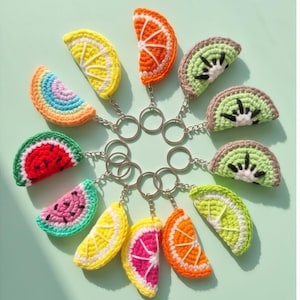 May include: A collection of crochet fruit slice keychains, including watermelon, lemon, orange, kiwi, and a rainbow-colored slice. Each keychain features a silver-colored ring and chain, and the fruit slices are in vibrant colors.