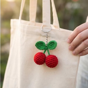 May include: A red and green crochet cherry keychain hanging from a canvas tote bag. The keychain features two red cherries with green leaves and a silver chain. The tote bag is a natural off-white color.