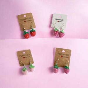 Könnte beinhalten: Vier Paar handgefertigte Erdbeer-Ohrringe. Die Ohrringe sind mit rotem, rosa und grünem Garn gehäkelt und an Ohrhaken befestigt. Jedes Paar ist auf einer Karte mit der Aufschrift "Handmade with love" befestigt. Der Hintergrund ist rosa.