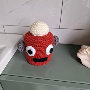 Puede incluir: Un personaje hecho a mano, tejido a ganchillo en rojo, con ojos blancos, boca negra y ap&eacute;ndices grises en forma de orejas. Una bola blanca est&aacute; encima de la cabeza del personaje. El art&iacute;culo est&aacute; sobre una superficie verde.