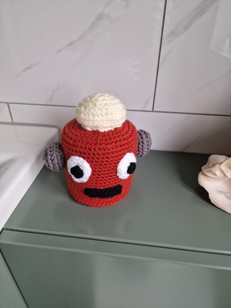 Puede incluir: Un personaje hecho a mano, tejido a ganchillo en rojo, con ojos blancos, boca negra y ap&eacute;ndices grises en forma de orejas. Una bola blanca est&aacute; encima de la cabeza del personaje. El art&iacute;culo est&aacute; sobre una superficie verde.