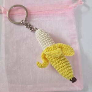 A crocheted banana keychain. The banana is yellow and white, with a small brown stem. It is attached to a silver metal key ring. The keychain is displayed on a pink organza bag.