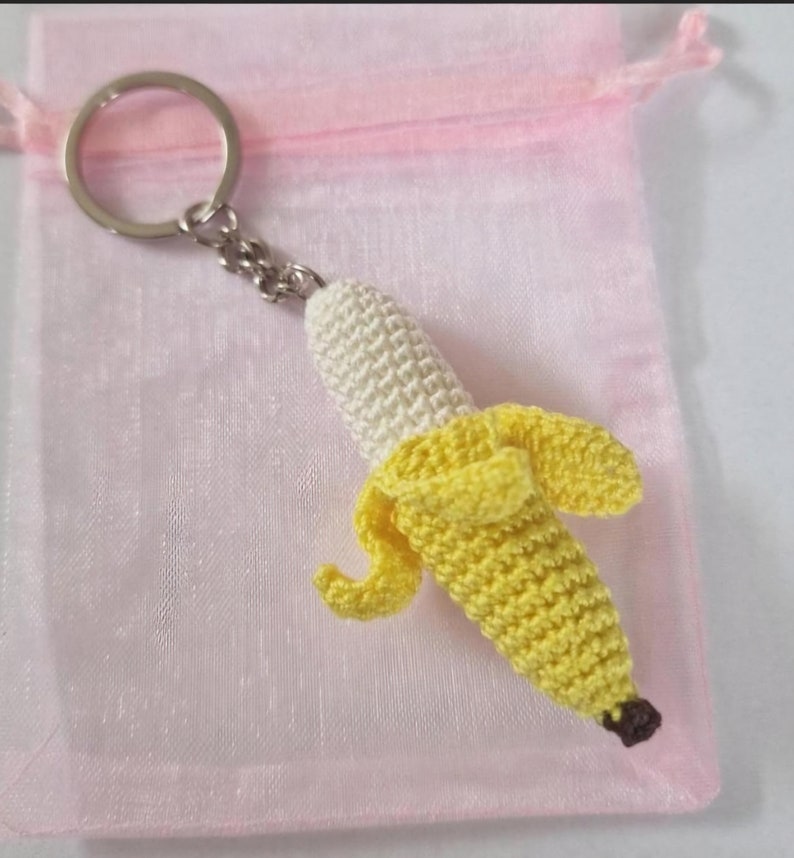 A crocheted banana keychain. The banana is yellow and white, with a small brown stem. It is attached to a silver metal key ring. The keychain is displayed on a pink organza bag.