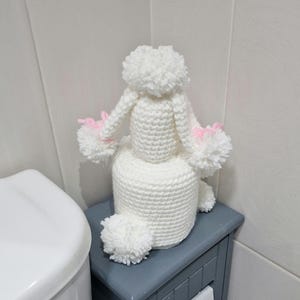 Puede incluir: Un portarrollos de papel higi&eacute;nico de caniche blanco tejido a ganchillo con detalles en rosa. El caniche est&aacute; sentado encima de un rollo de papel higi&eacute;nico. El soporte est&aacute; sobre un armario gris junto a un inodoro blanco.