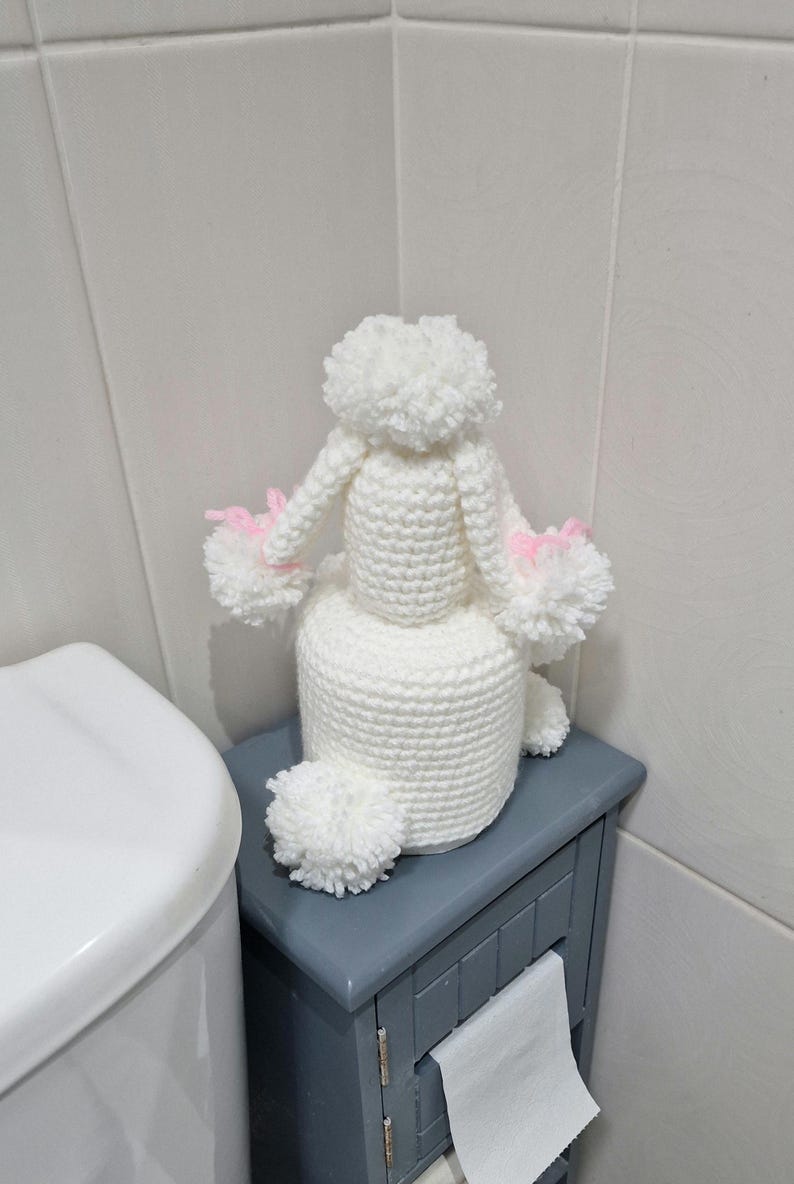 Puede incluir: Un portarrollos de papel higi&eacute;nico de caniche blanco tejido a ganchillo con detalles en rosa. El caniche est&aacute; sentado encima de un rollo de papel higi&eacute;nico. El soporte est&aacute; sobre un armario gris junto a un inodoro blanco.